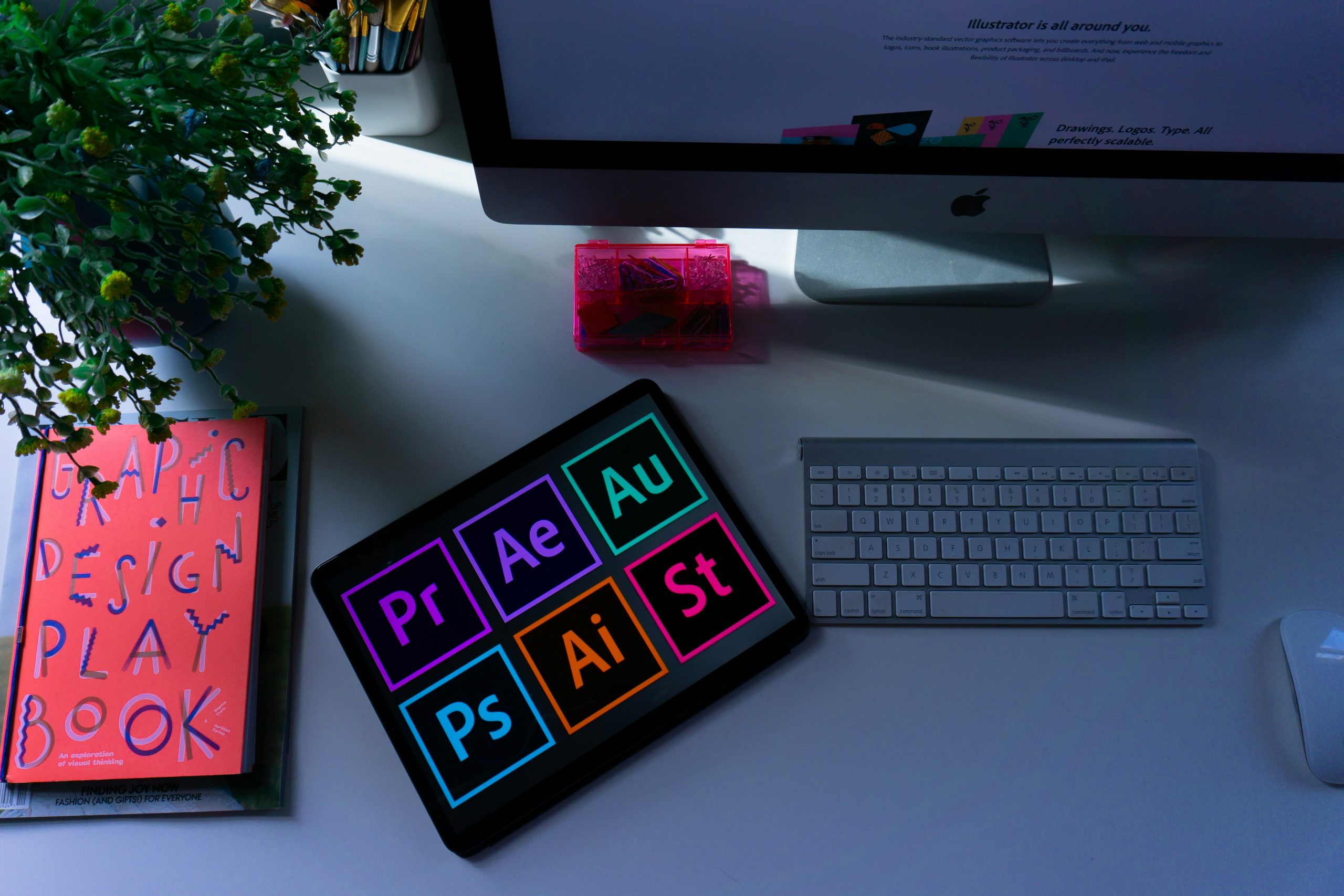 Professional workspace featuring a tablet displaying Adobe Premiere, After Effects, and Photoshop icons for creative design services.